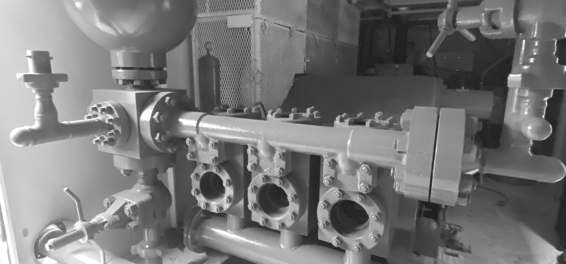 The fluid end of a triplex pump in a white enclosure, three bolted valve covers in a row under the discharge manifold