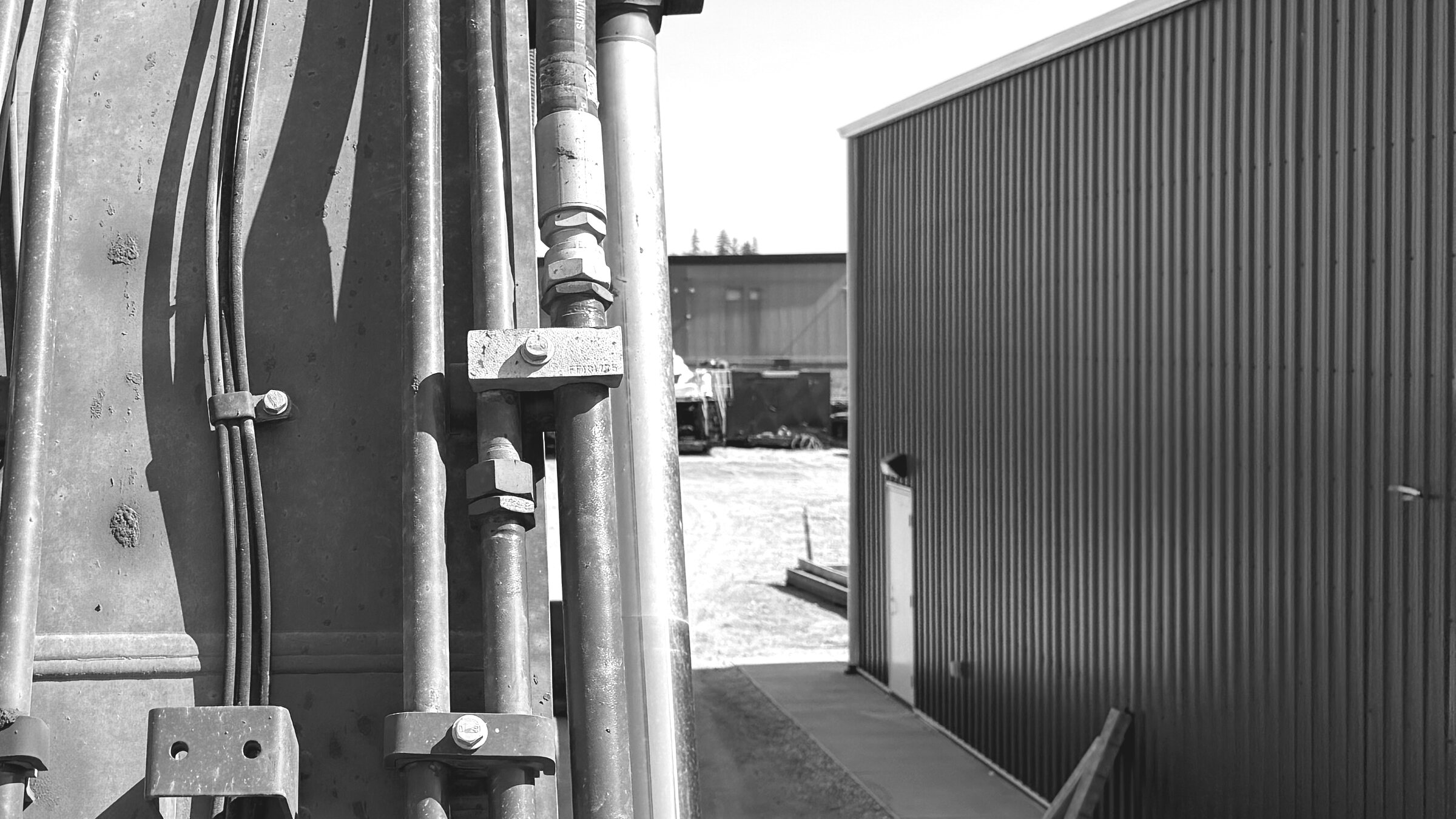 A dozer blade's hydraulic cylinders and hose clamps beside a corrugated-steel shop wall